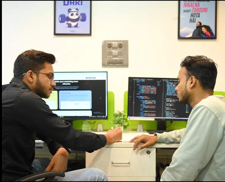 Two digital marketing professionals discussing website development and coding work in a modern office setup with dual monitors.